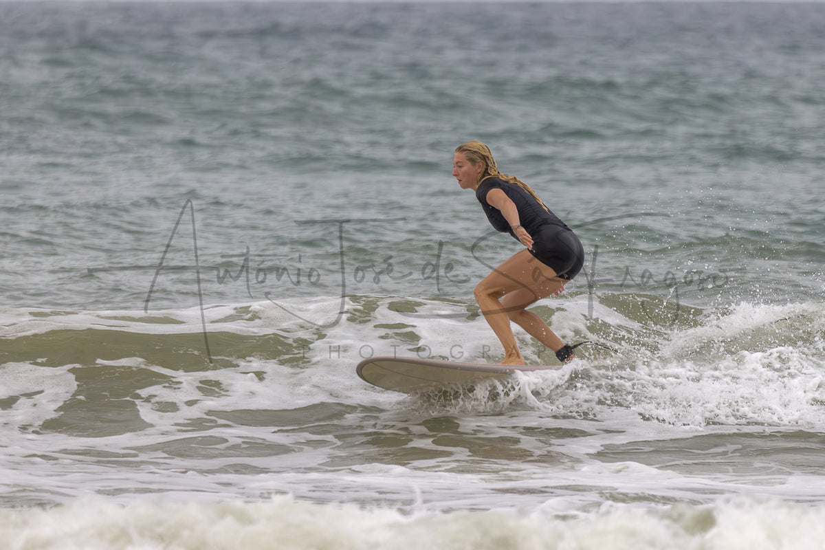 A woman in a black wetsuit stands on a surfboard in the ocean waves, with a large photographer watermark across the center.