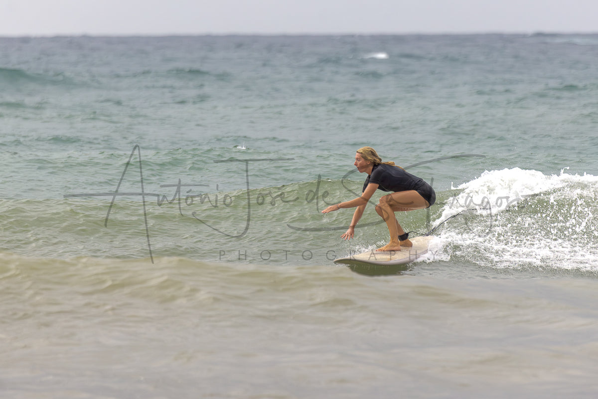A surfer in a black wetsuit crouches on a white surfboard on a small ocean wave, with the sea and cloudy sky in the background. A large photographer watermark is visible across the center.