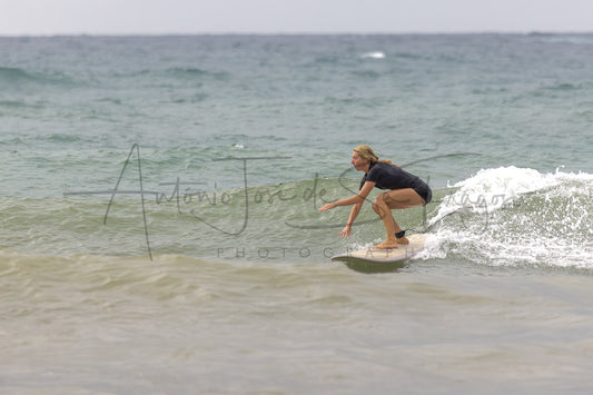 A surfer in a black wetsuit crouches on a white surfboard on a small ocean wave, with the sea and cloudy sky in the background. A large photographer watermark is visible across the center.