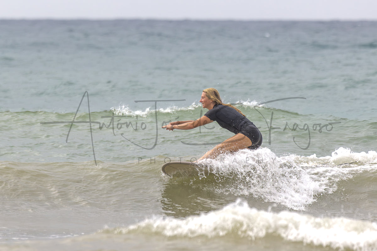 A woman in a black wetsuit rides a surfboard on a small ocean wave, with the sea and cloudy sky in the background. A large semi-transparent photographer watermark is visible across the center.
