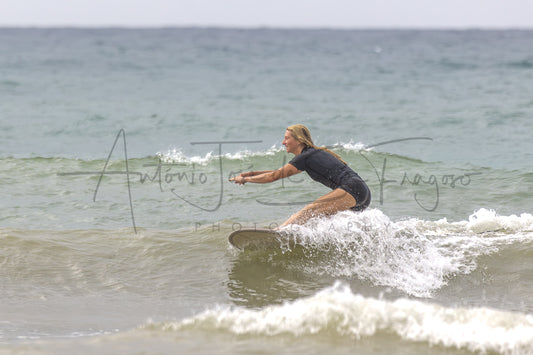 A woman in a black wetsuit rides a surfboard on a small ocean wave, with the sea and cloudy sky in the background. A large semi-transparent photographer watermark is visible across the center.