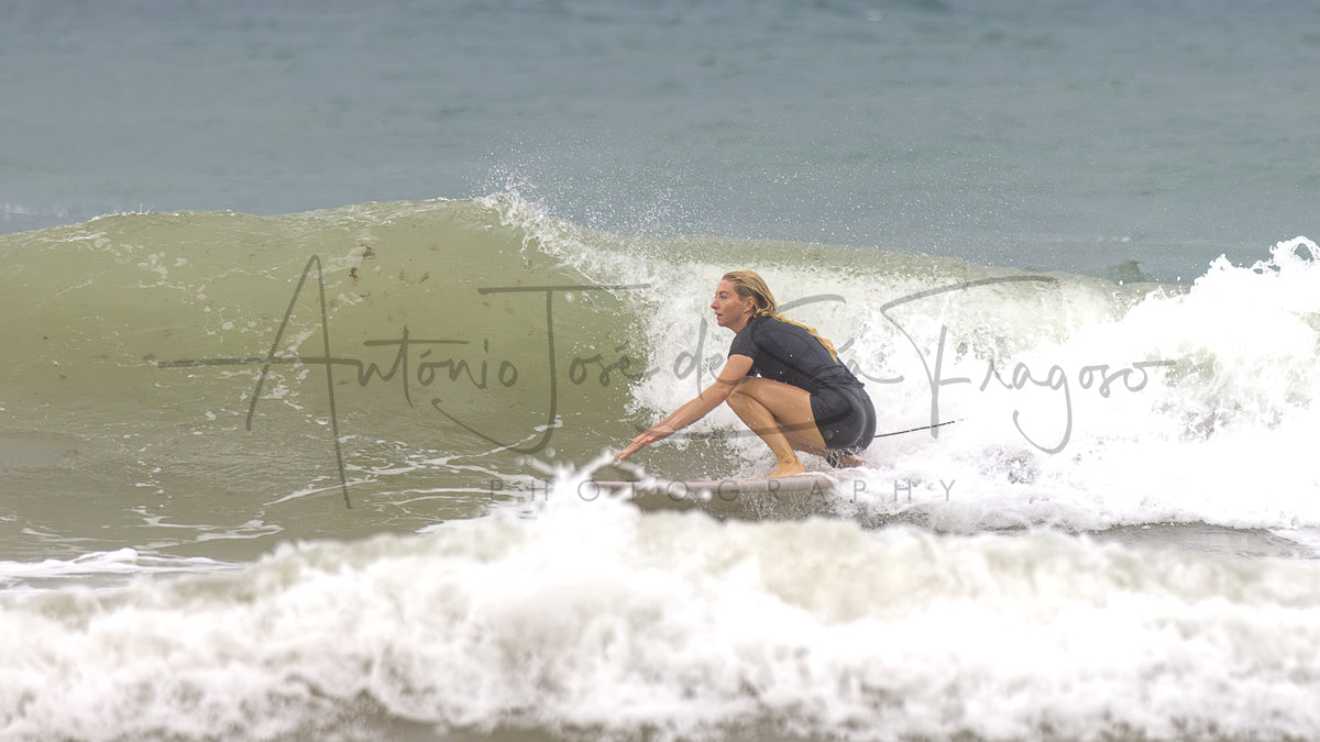A woman in a black wetsuit crouches on a surfboard riding a small wave in the ocean, with white sea foam in the foreground and a large translucent watermark across the center.