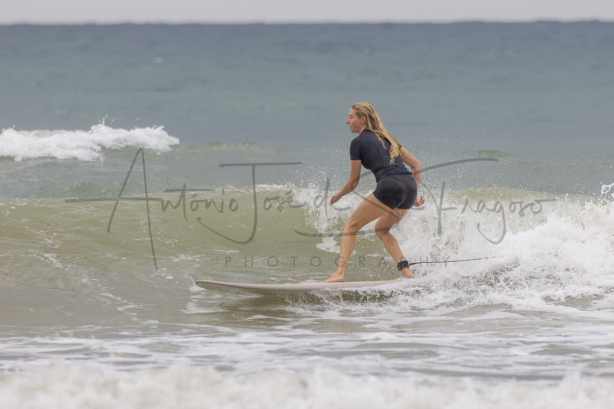 A woman in a black wetsuit rides a surfboard on a gentle ocean wave, with white sea foam and a cloudy sky in the background. A large photographer watermark is visible across the center.