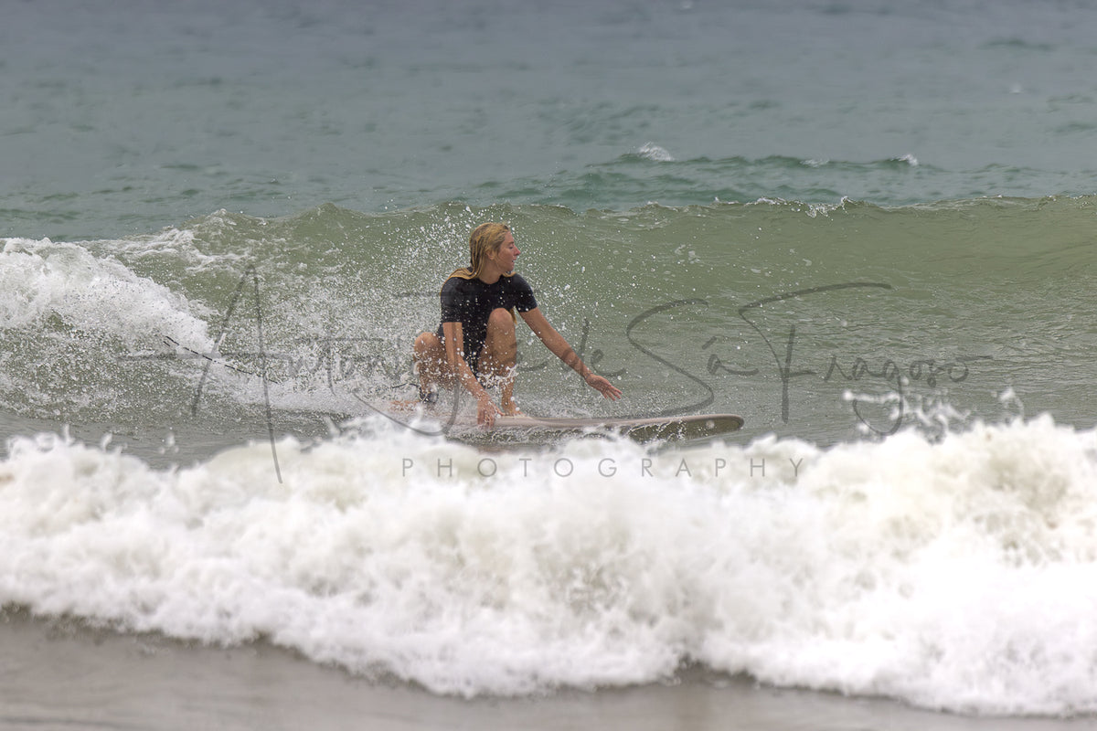 A surfer rides a small wave on a surfboard at the beach, wearing a black swimsuit. White sea foam surrounds the surfer, with blue-gray ocean water in the background.