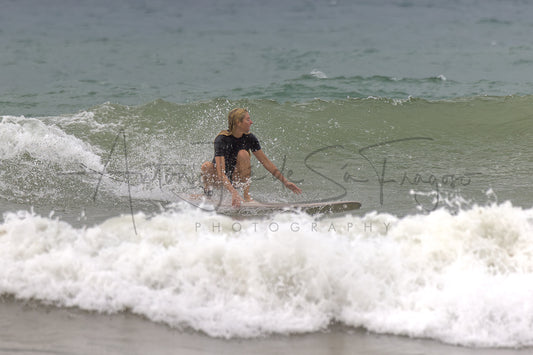 A surfer rides a small wave on a surfboard at the beach, wearing a black swimsuit. White sea foam surrounds the surfer, with blue-gray ocean water in the background.