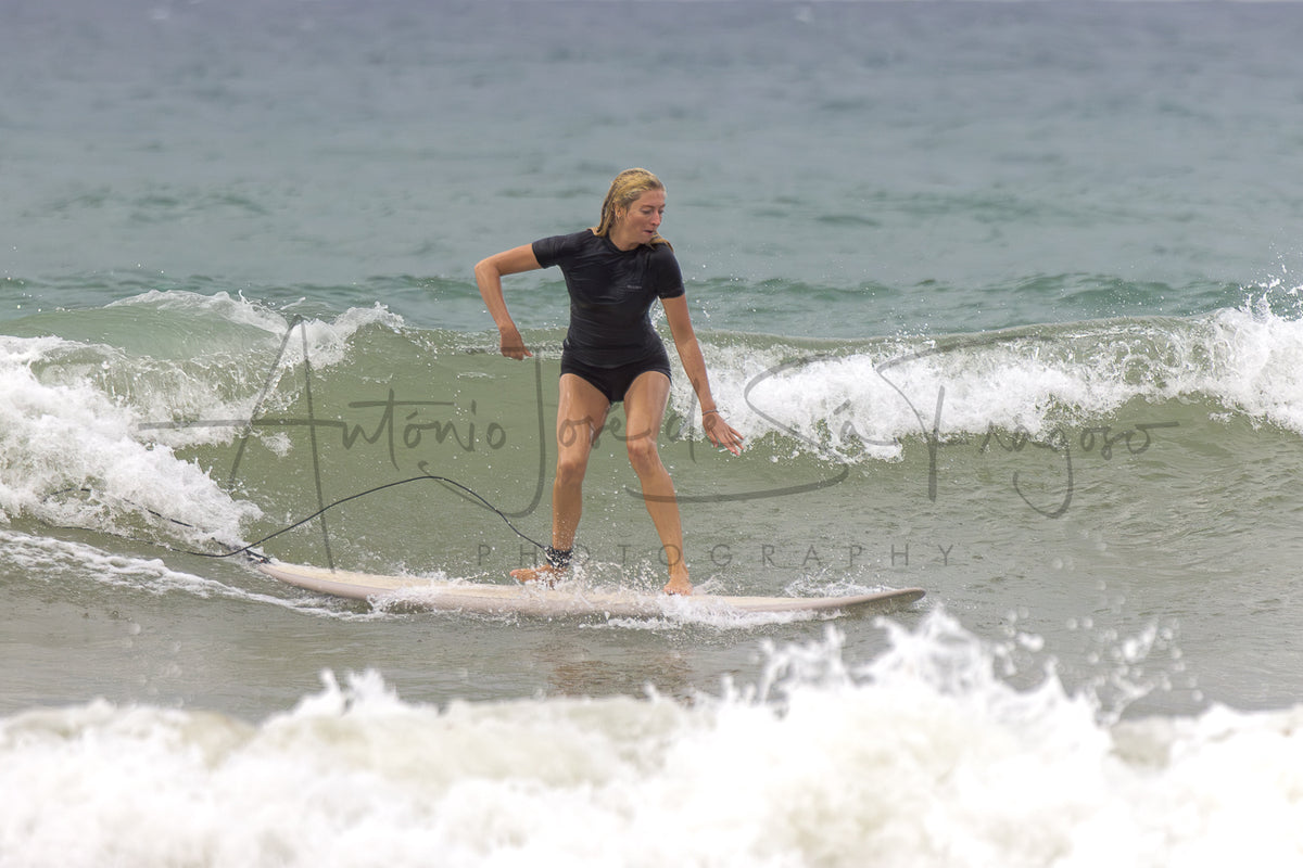 A woman in a black wetsuit stands on a surfboard on a gentle ocean wave, with large overlaid text reading 'António José de Sá Fragoso PHOTOGRAPHY'.