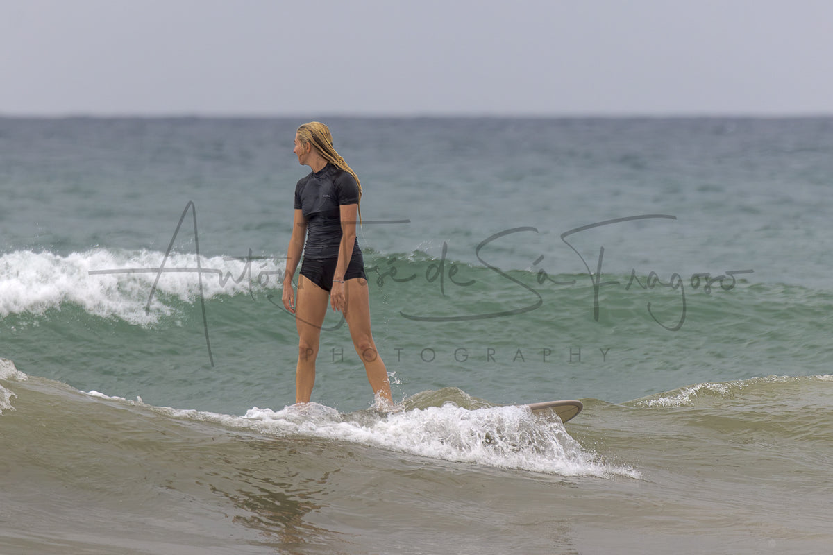 A woman in a black wetsuit stands on a surfboard in the ocean waves, with a large black photographer's watermark across the center of the image.