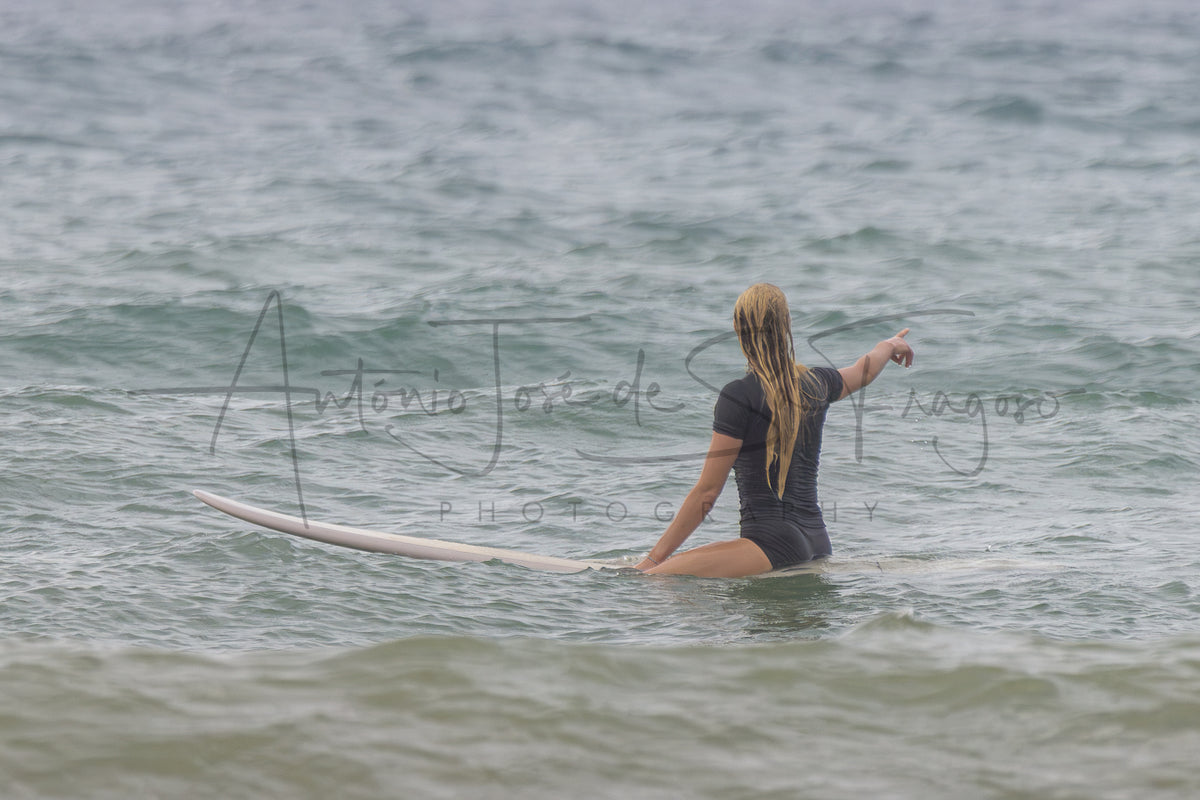 A woman in a dark gray swimsuit sits on a white surfboard in the ocean, viewed from behind. A large black script watermark reading 'Antônio José de Souza Photography' is overlaid across the center.