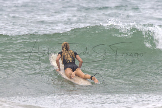 A surfer wearing a black one-piece swimsuit lies on a white surfboard in the ocean, paddling behind a breaking green wave. A large semi-transparent watermark reading 'Antonio de San Fragaso' is overlaid across the center.