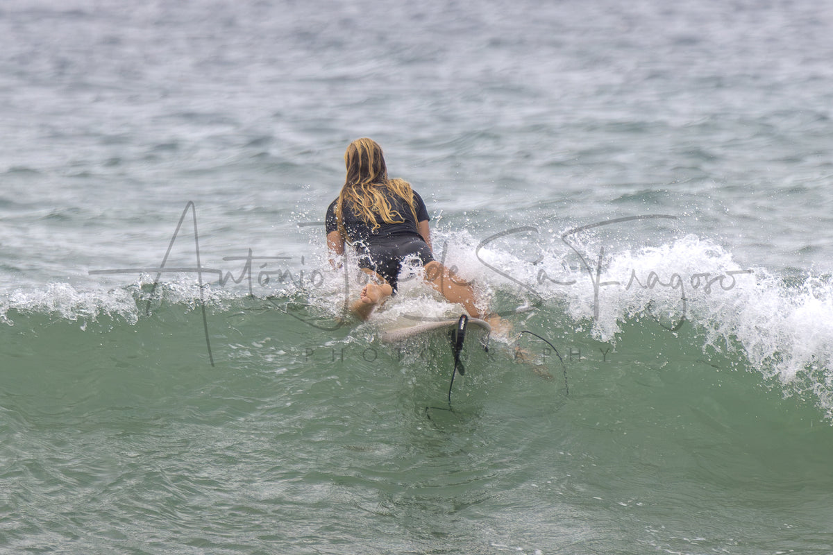 A surfer with long blonde hair, seen from behind, riding a small wave in the ocean. The surfer is wearing a black swimsuit.