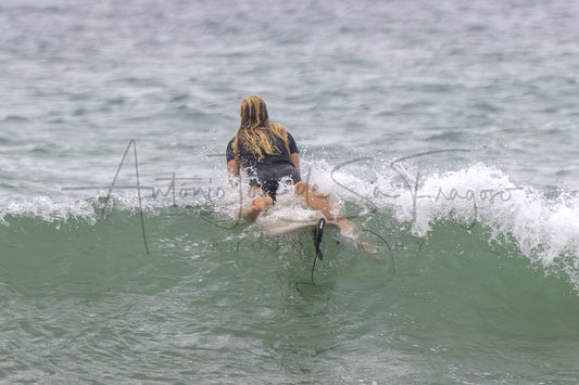 A surfer with long blonde hair, seen from behind, riding a small wave in the ocean. The surfer is wearing a black swimsuit.
