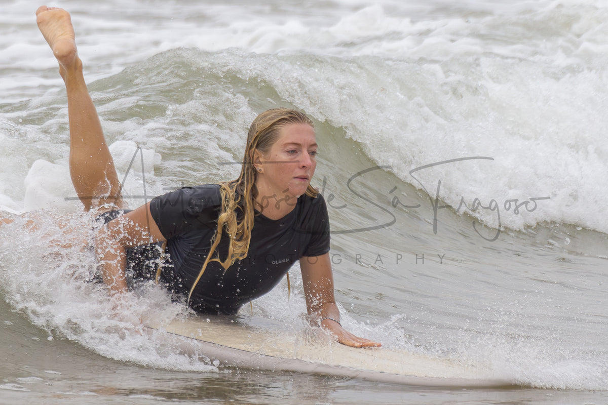 A woman in a black wetsuit lies on a surfboard paddling in a breaking ocean wave, with a large script watermark across the center reading 'Antonio José de Sá Fragoso'.
