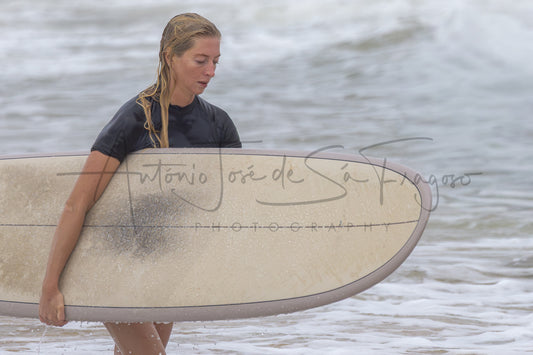 A woman in a dark t-shirt carrying a surfboard in the ocean water, with a large photographer's watermark across the center.