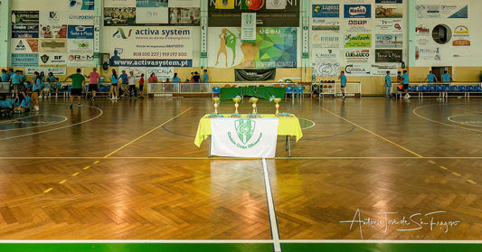Indoor sports hall with a table and chairs setup, surrounded by various advertisements on the walls.