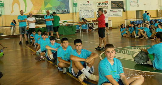 Group of young people in blue shirts sitting on a gymnasium floor with sports equipment and banners in the background.