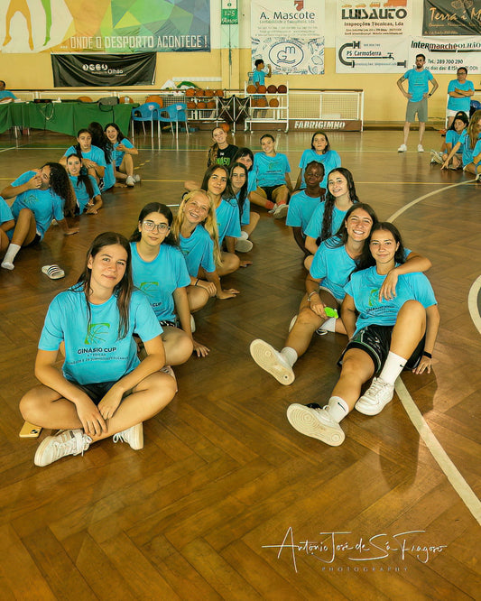 Group of people in blue shirts sitting on a wooden floor in a sports hall.