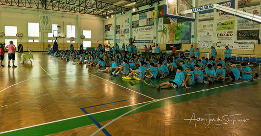 Children in blue shirts sitting on the floor of a gymnasium with sports equipment and banners.