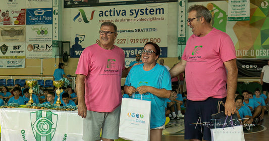 Two men in pink shirts standing with a woman in a blue shirt holding a white bag, with a sports arena in the background.