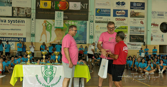 Two men in pink shirts standing next to a table with trophies in a sports hall.