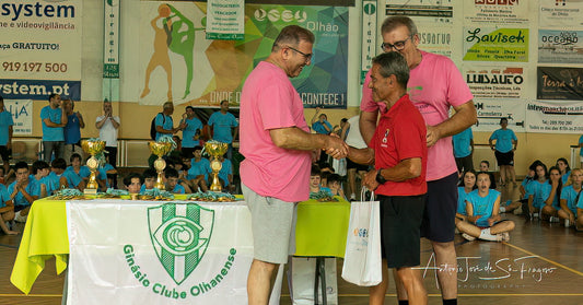 Two men in pink shirts shaking hands at a sports event with trophies and children in the background.