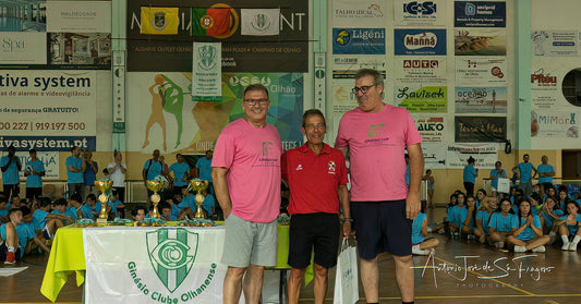 Three men in pink shirts standing in front of a banner with a sports club logo, surrounded by children and advertisements on the wall.