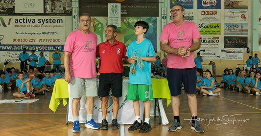Three men standing on a podium with children in a sports hall, surrounded by spectators and advertisements.