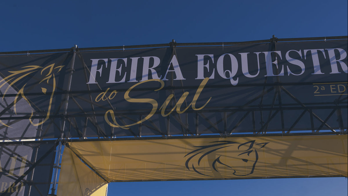 A large outdoor banner and archway for 'Feira Equestria do Sul' with white and gold lettering on a dark blue fabric, supported by a metal truss structure against a clear blue sky.