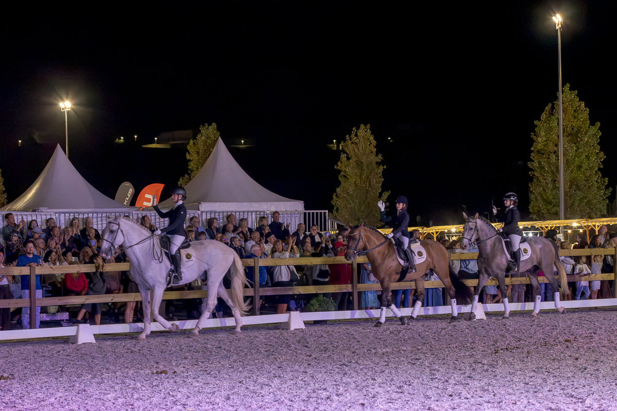 Three horse riders on horseback compete in a night equestrian event, with a crowd of spectators behind a fence and tents in the background under stadium lights.