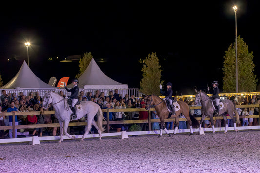 Three horse riders on horseback compete in a night equestrian event, with a crowd of spectators behind a fence and tents in the background under stadium lights.