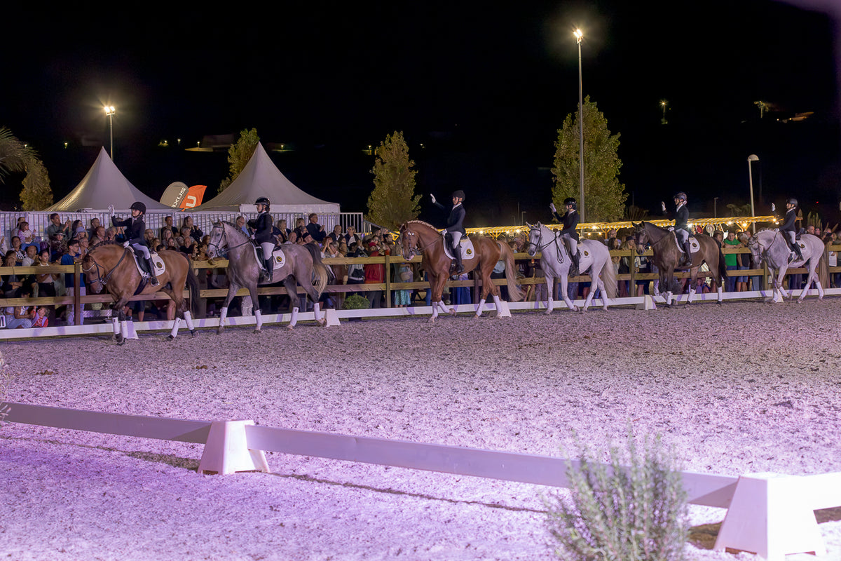 A night equestrian event scene showing several riders on horseback in an arena, with spectators behind barriers and white tents in the background under stadium lights.