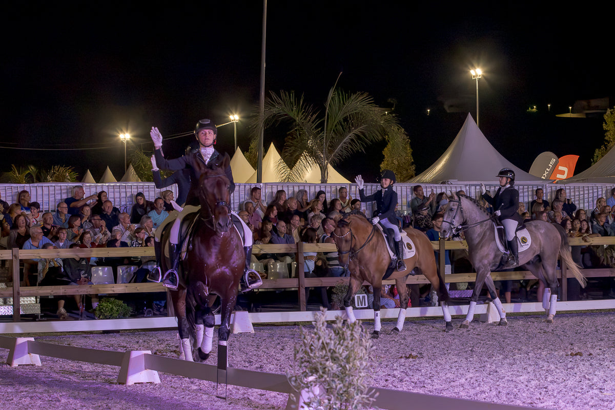 A night-time equestrian event scene showing three riders on horseback in an arena, with a crowd of spectators behind wooden barriers and tents in the background under bright stadium lights.