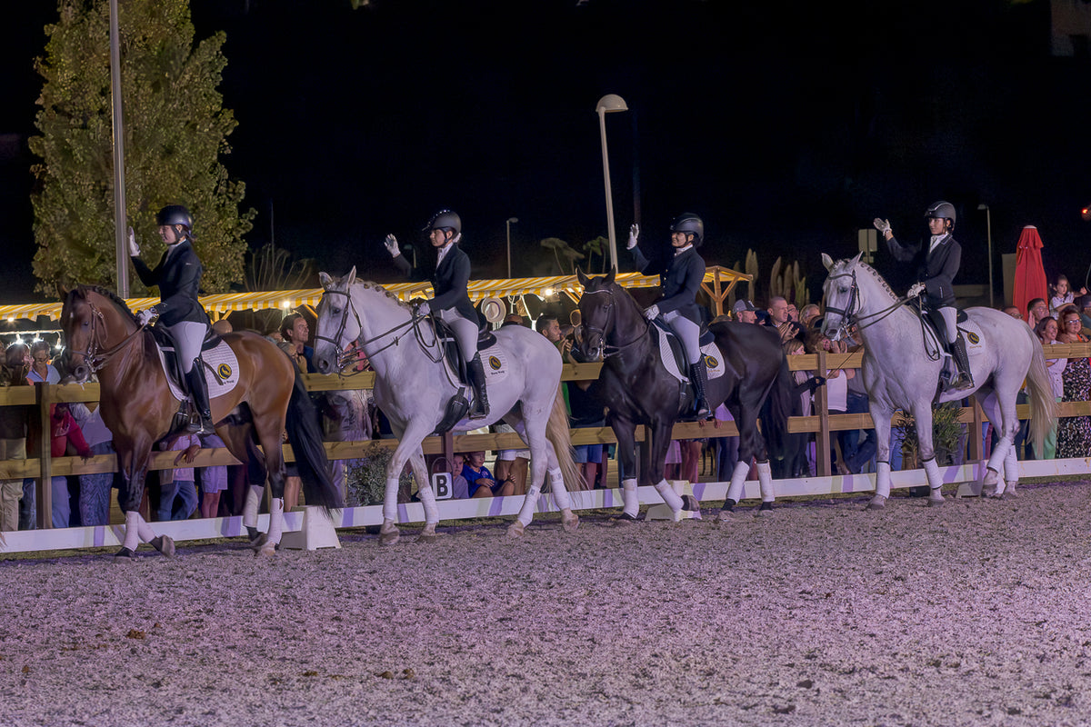 Four equestrians on horseback ride in a nighttime event at an arena, with spectators visible behind a fence in the background.