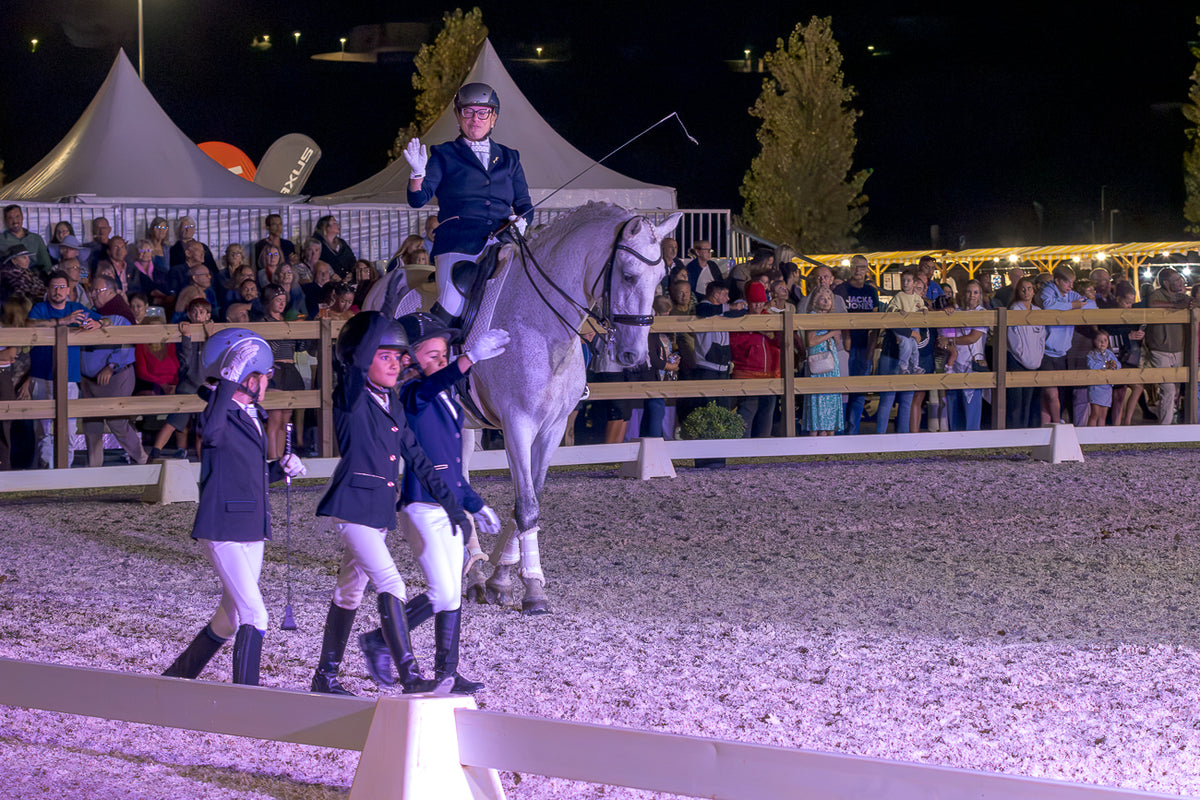 Equestrian riders in formal attire on a horse and on foot inside an outdoor riding arena at night, with spectators standing behind a fence under bright event lighting.