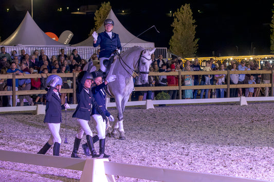 Equestrian riders in formal attire on a horse and on foot inside an outdoor riding arena at night, with spectators standing behind a fence under bright event lighting.