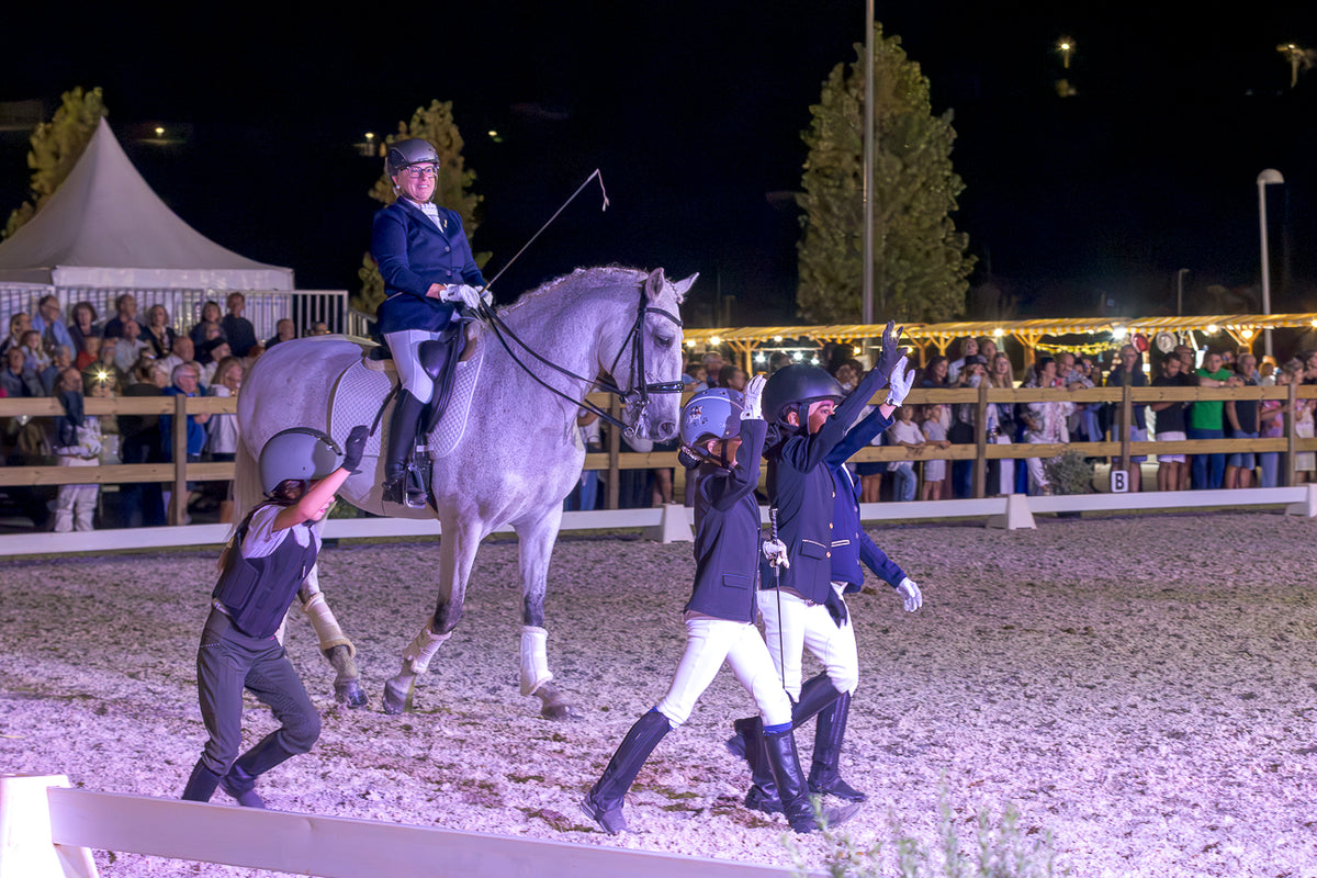 A night equestrian event in an arena with a rider on a light gray horse, surrounded by handlers and spectators behind a fence under bright lights.