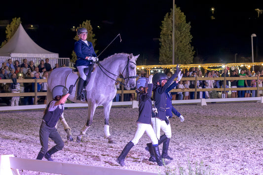 A night equestrian event in an arena with a rider on a light gray horse, surrounded by handlers and spectators behind a fence under bright lights.