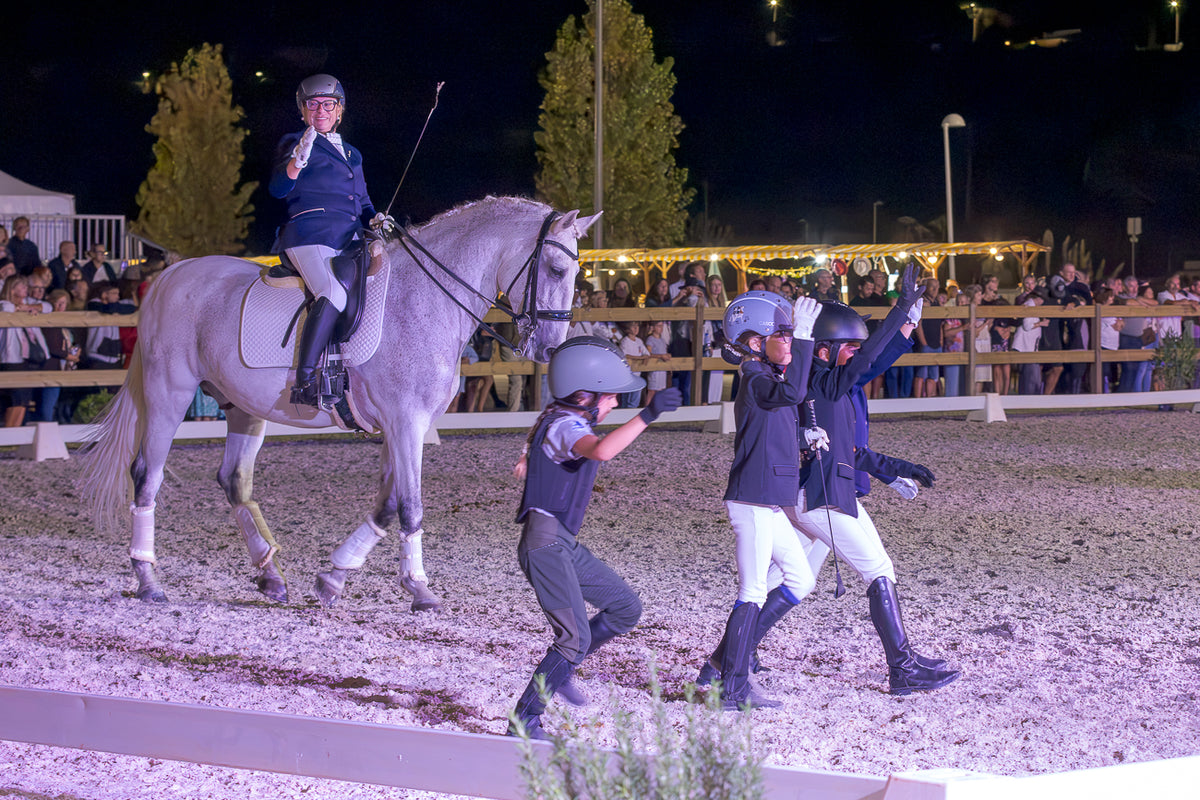 A night-time equestrian scene in an arena with a rider on a light gray horse, surrounded by riders and spectators behind a fence.