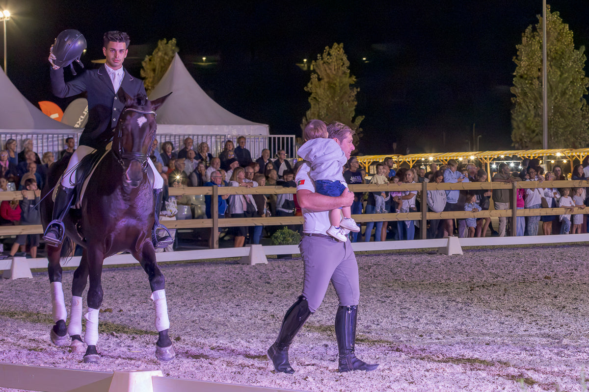 A night-time equestrian event scene in an arena, showing a rider in dark formalwear on a dark horse while another person in riding apparel carries a child near the center, with spectators and tents in the background.