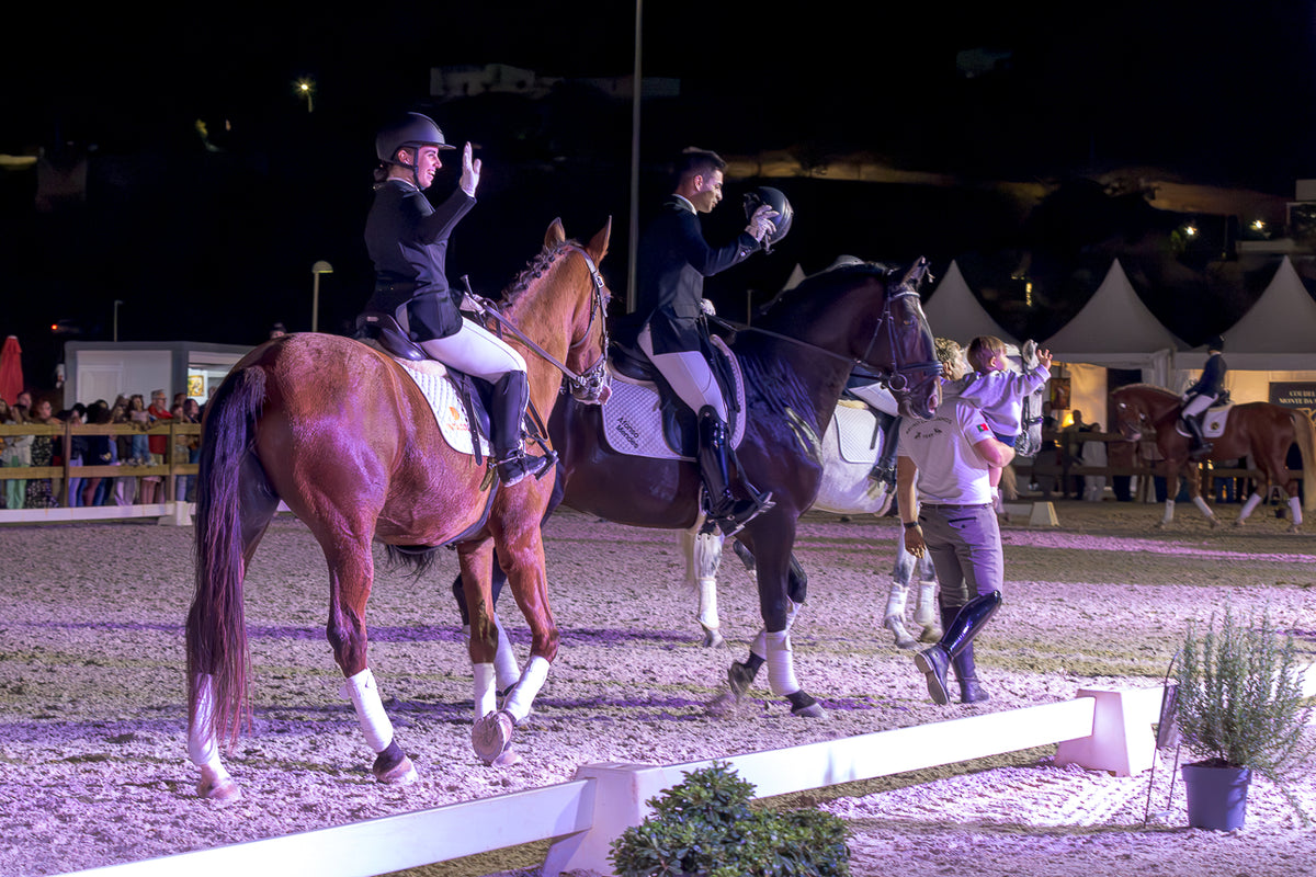 Two equestrian riders on horseback at night in an outdoor riding arena, with a handler walking beside them. The horses are one chestnut brown and one dark bay, both wearing white leg wraps, under event lighting.