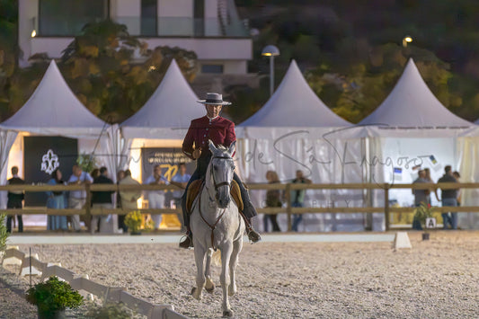 A person in a portuguese traditional hat and dark outfit rides a white horse across a sand arena at night, with white event tents and spectators visible in the background.