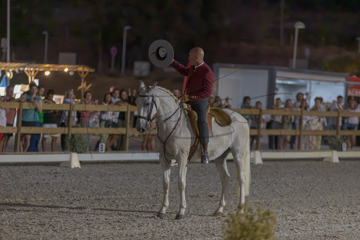 A man in a red shirt and dark pants rides a white horse in an outdoor arena at night while holding up a large circular hat. Spectators are visible behind wooden fences in the background.