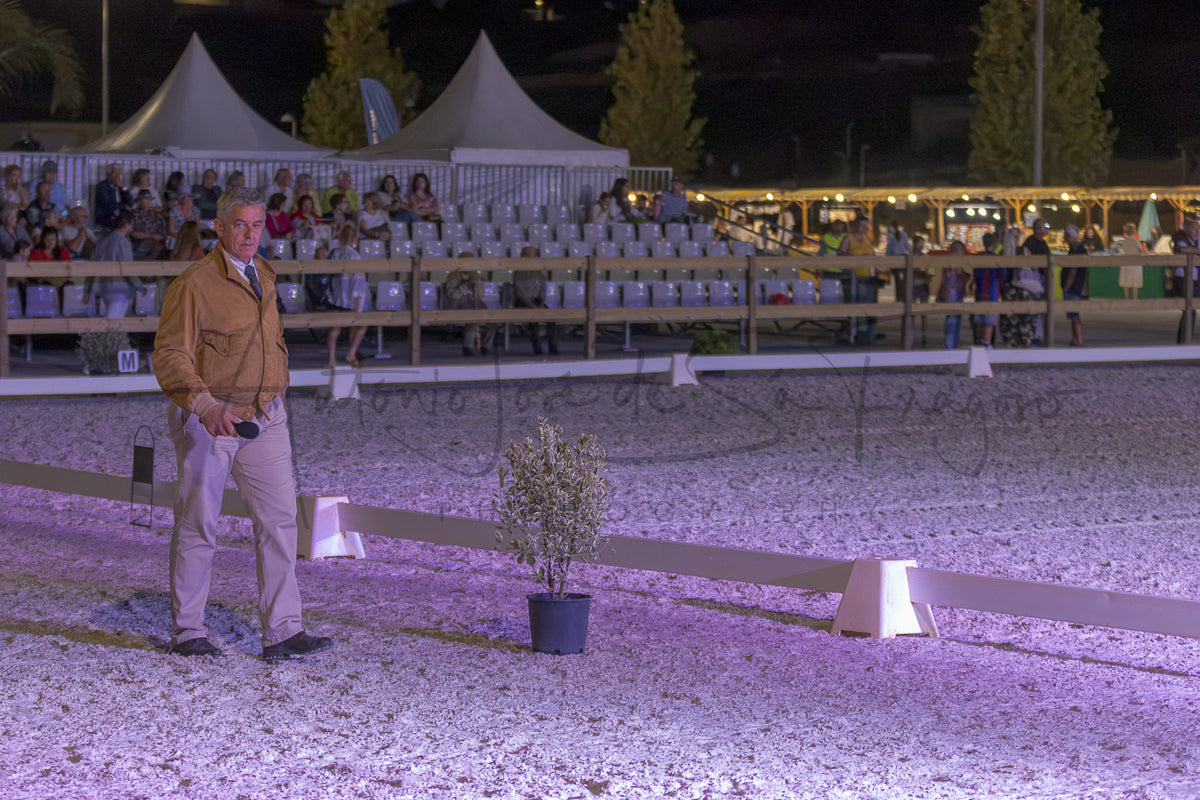 A man in a tan jacket and khaki pants stands in a gravel-covered arena at night under event lighting, with a potted plant in the foreground and spectators seated in tents in the background.