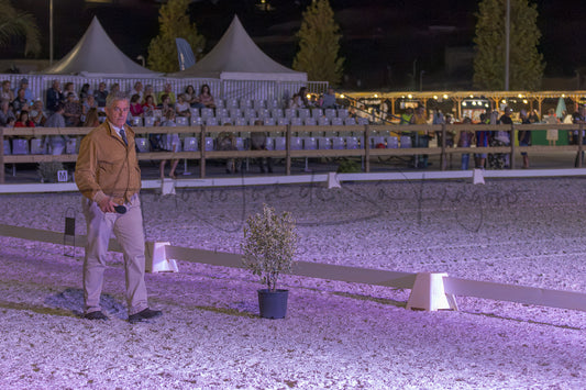 A man in a tan jacket and khaki pants stands in a gravel-covered arena at night under event lighting, with a potted plant in the foreground and spectators seated in tents in the background.