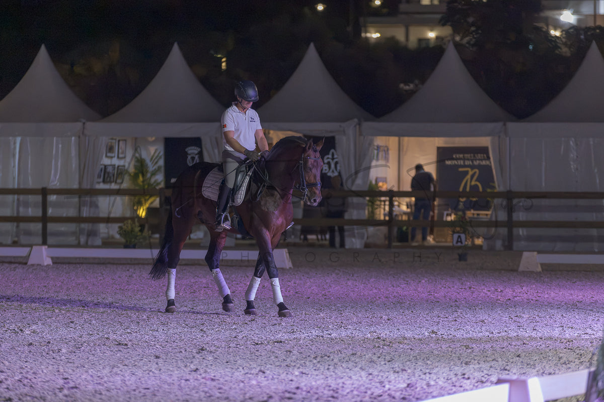 A rider on a dark brown horse walks in an equestrian arena at night under artificial lighting, with white event tents and fencing in the background.