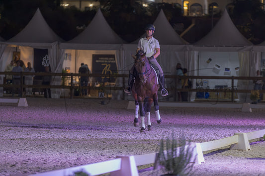 A horse rider on a brown horse in an equestrian arena at night, with white event tents and spectators visible in the background.