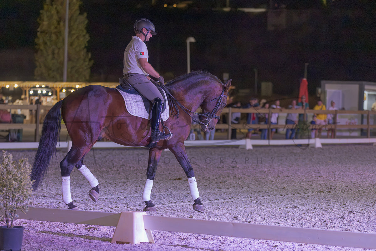 A rider in equestrian attire sits on a dark brown horse with white leg wraps inside an outdoor riding arena at night under artificial lighting.