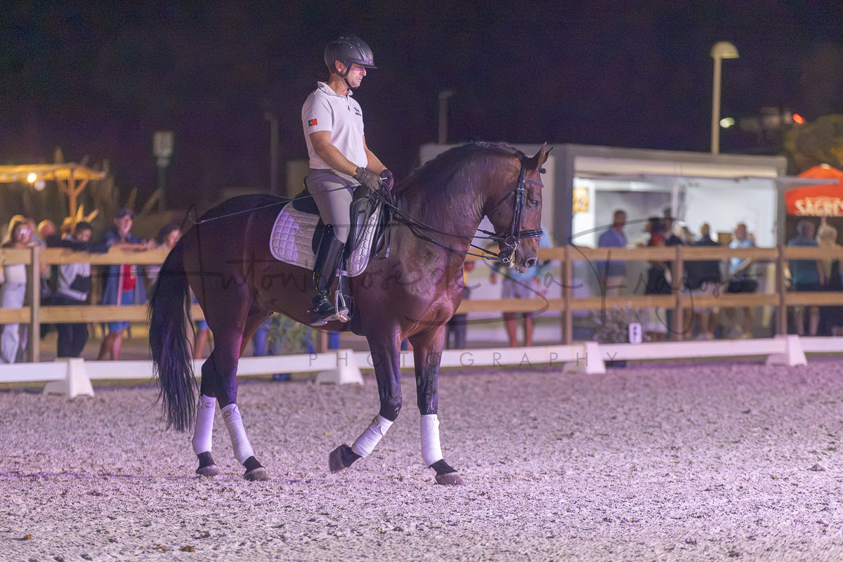 A dark brown horse with white leg wraps is being ridden by a person in equestrian attire inside an outdoor riding arena at night.
