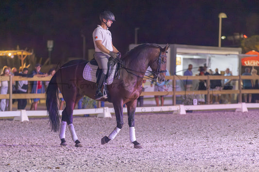 A dark brown horse with white leg wraps is being ridden by a person in equestrian attire inside an outdoor riding arena at night.