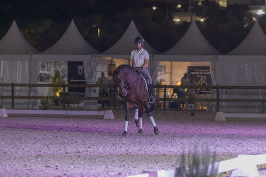 A rider on a dark brown horse performs a jump in an equestrian arena at night, with white event tents and fencing visible in the background.