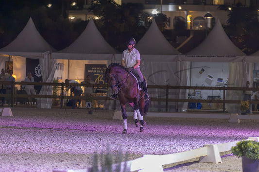 A horse rider on a dark brown horse during an evening equestrian event, with white event tents and a lit arena in the background.
